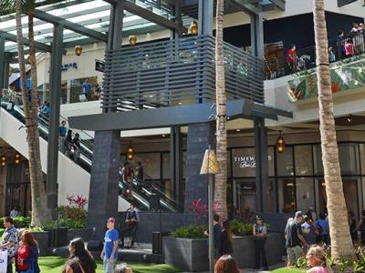 People walking around outdoors at Ala Moana shopping center on a sunny day. Bright green grass in the foreground. An escalator to second floor shows in the background.